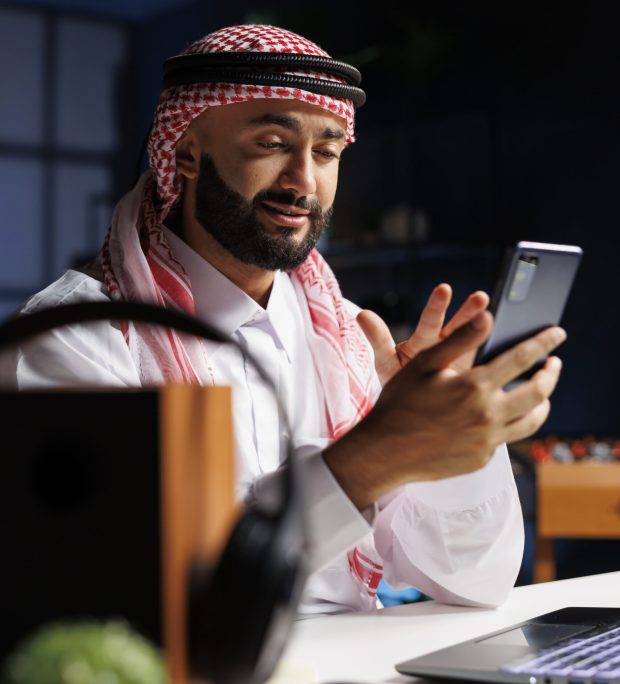 Middle Eastern entrepreneur having a serious conversation with a coworker on his mobile device in his home office. He wears Islamic clothing and has a professional demeanor.